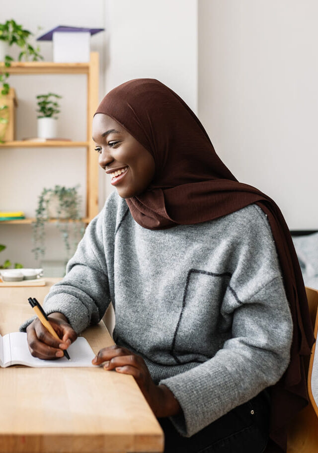 A woman wearing a hijab sits at a desk, focused on her laptop.