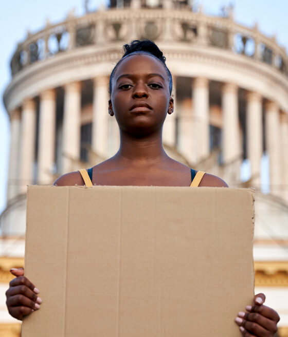 A young woman holding a cardboard sign in front of a building, possibly protesting or seeking help.